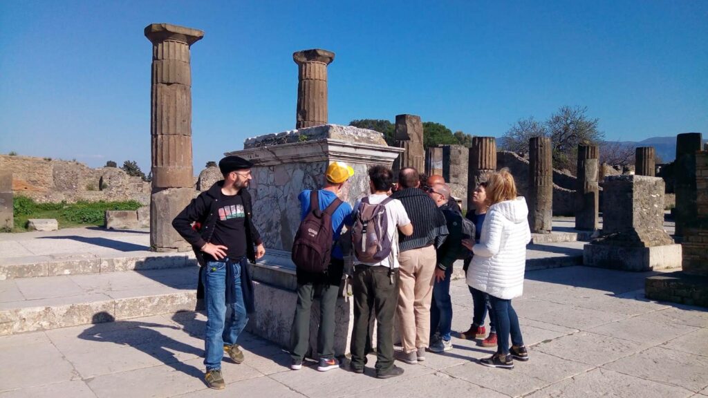 Un gruppo di viaggiatori a Pompei, riuniti intorno ad un piedistallo nel foro principale. Dietro di loro alcune colonne ed i muri di antiche case, sopra di loro il cielo azzurro.