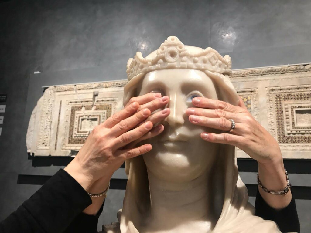 Tre mani toccano gli occhi di vetro di una replica di Madonna trecentesca in pietra bianca al Museo dell'Opera del Duomo.