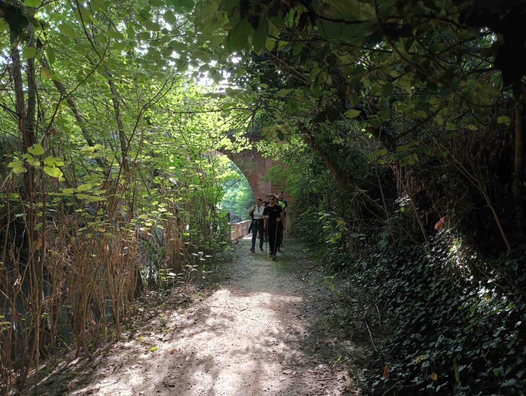 Un gruppo di camminatori, al centro dell'immagine, procede lungo un sentiero circondato da sottobosco fluviale. Dietro di loro, tra le foglie si intravede l'arco in mattoni di un ponte.