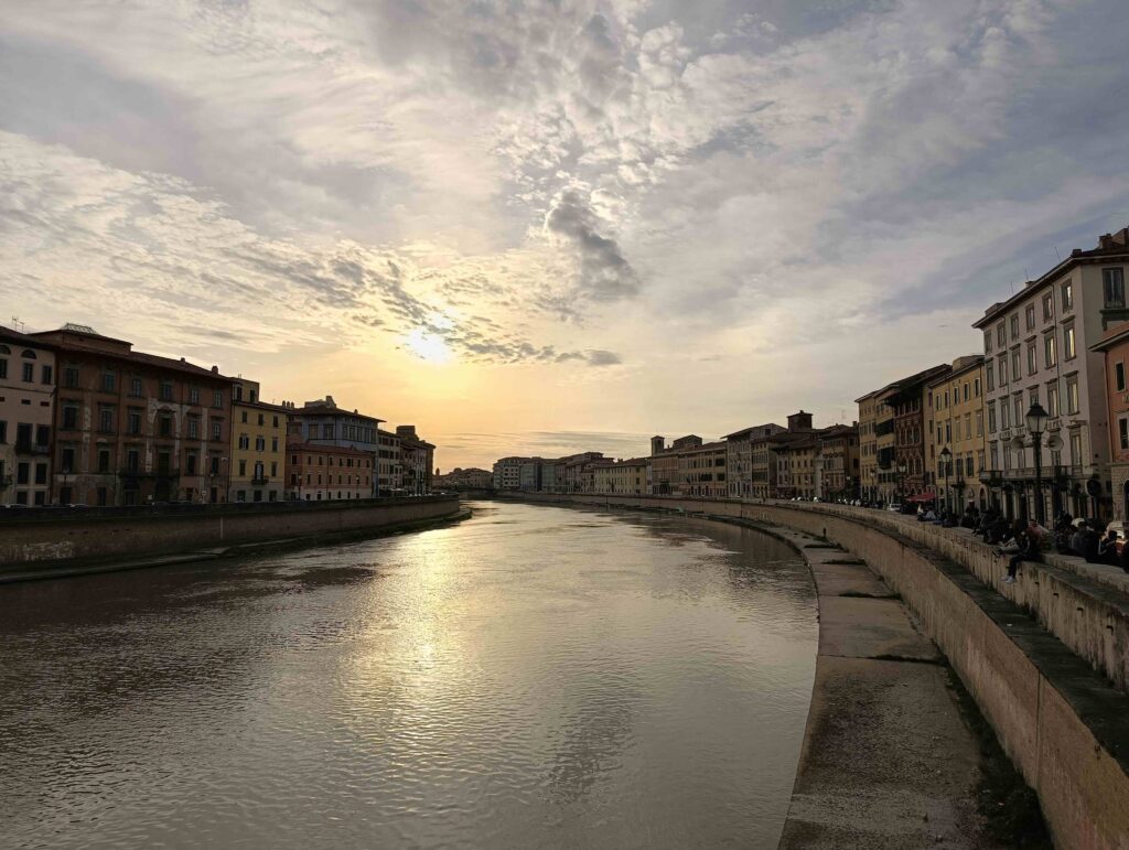 Il fiume Arno a Pisa che piega verso sinistra guardando verso il tramonto, nel centro della città, con cortine di palazzi semplici ma eleganti lungo entrambe le rive.