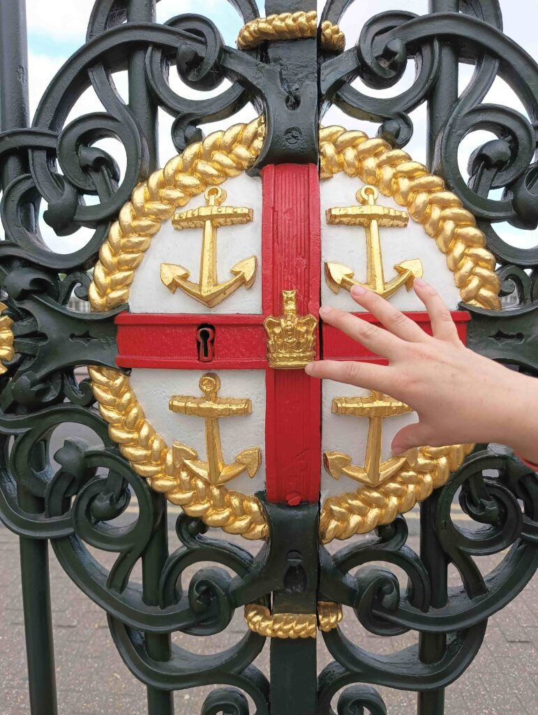 Una mano tocca il simbolo del Royal Naval College a Londra in ferro battuto su un cancello: una croce di San Giorgio - rossa su fondo bianco - all'interno di un cerchio delimitato da una corda dorata, con al centro una corona dorata e nei quattro quadranti delle ancore, pure dorate.