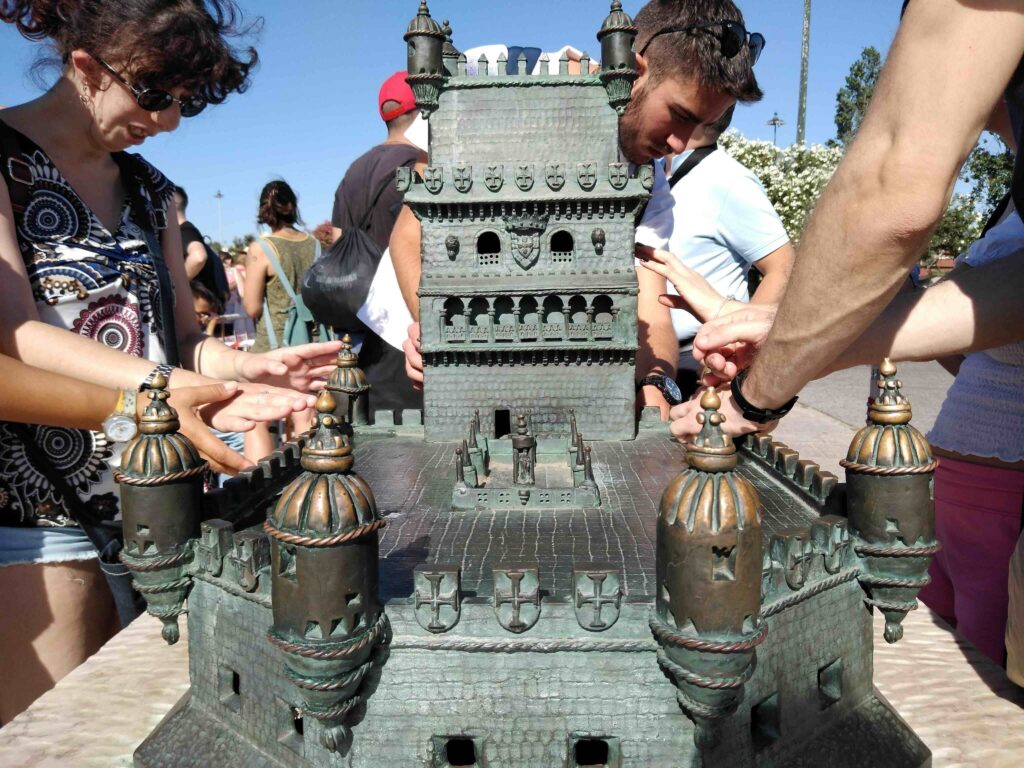 Diverse persone toccano un modellino di bronzo della cinquecentesca Torre di Belém, raffigurato in primo piano.