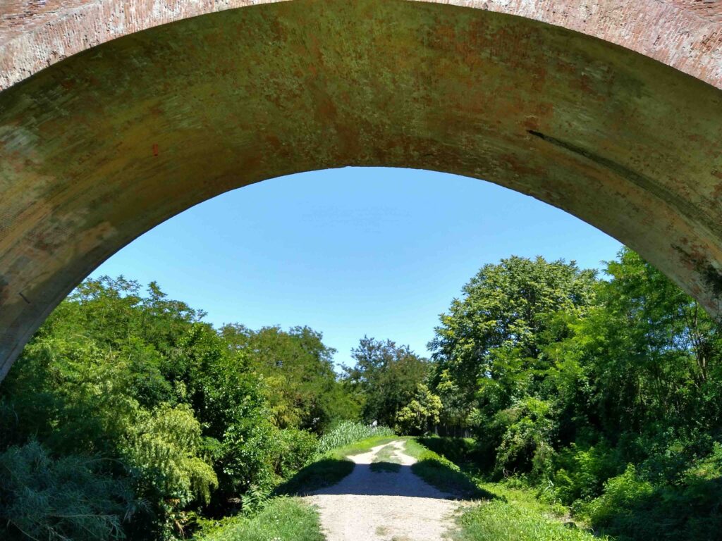 Un sentiero tra due canali - quello a destra, più grande, è il Navile - si snoda nella distanza sotto all'arcata in mattoni di un ponte.