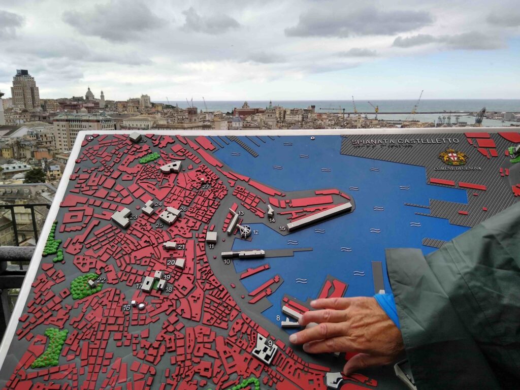 In primo piano, una mano tocca un pannello a rilievo raffigurante il centro storico di Genova. Dietro al pannello, il panorama si apre sui tetti di Genova, il porto, il mare e l'orizzonte sotto un cielo annuvolato.