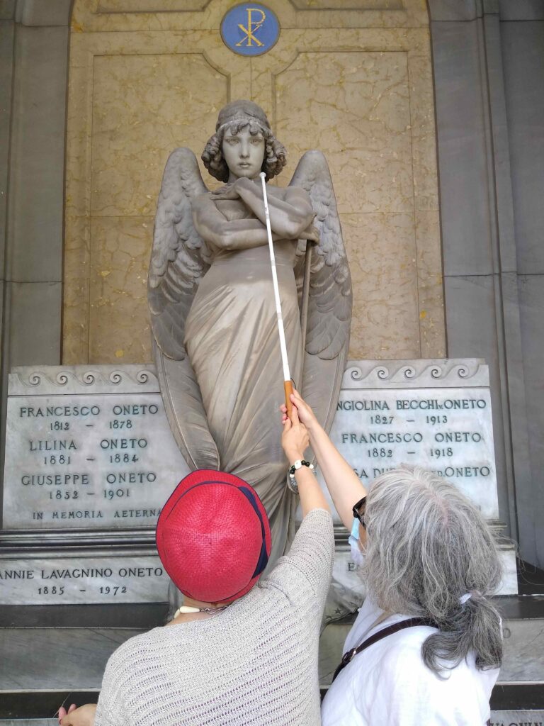 Due persone di spalle: una guida l'altra nell'esplorazione col bastone bianco di una statua funebre raffigurante un angelo, posta in posizione sopraelevata, al cimitero monumentale di Staglieno.