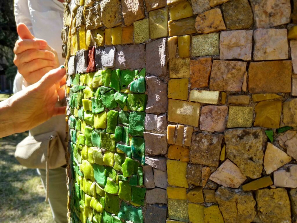 Mani toccano tessere di mosaico irregolari verdi e gialle, sia di vetro che di pietra, che rivestono una scultura contemporanea di forma cilindrica.