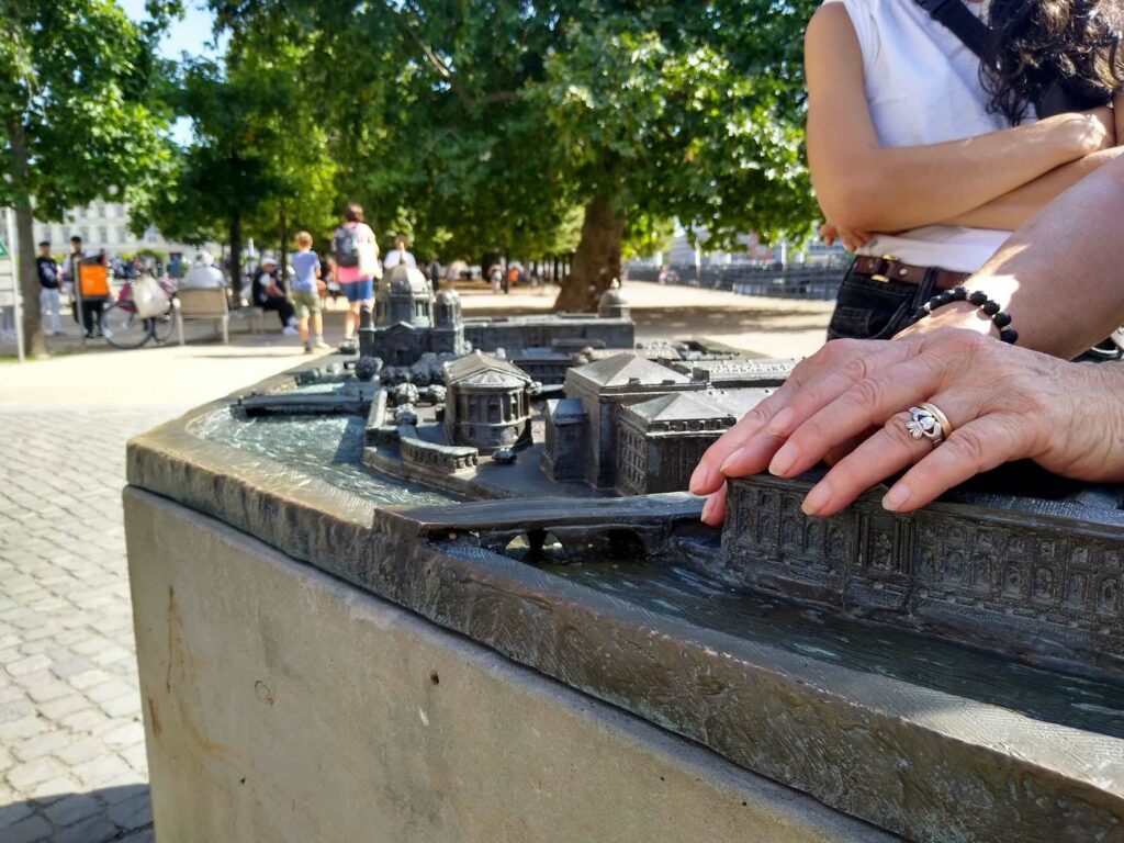 Sul fondo di giornata di sole, mani in primo piano toccano un plastico in bronzo che raffigura l'Isola dei Musei.