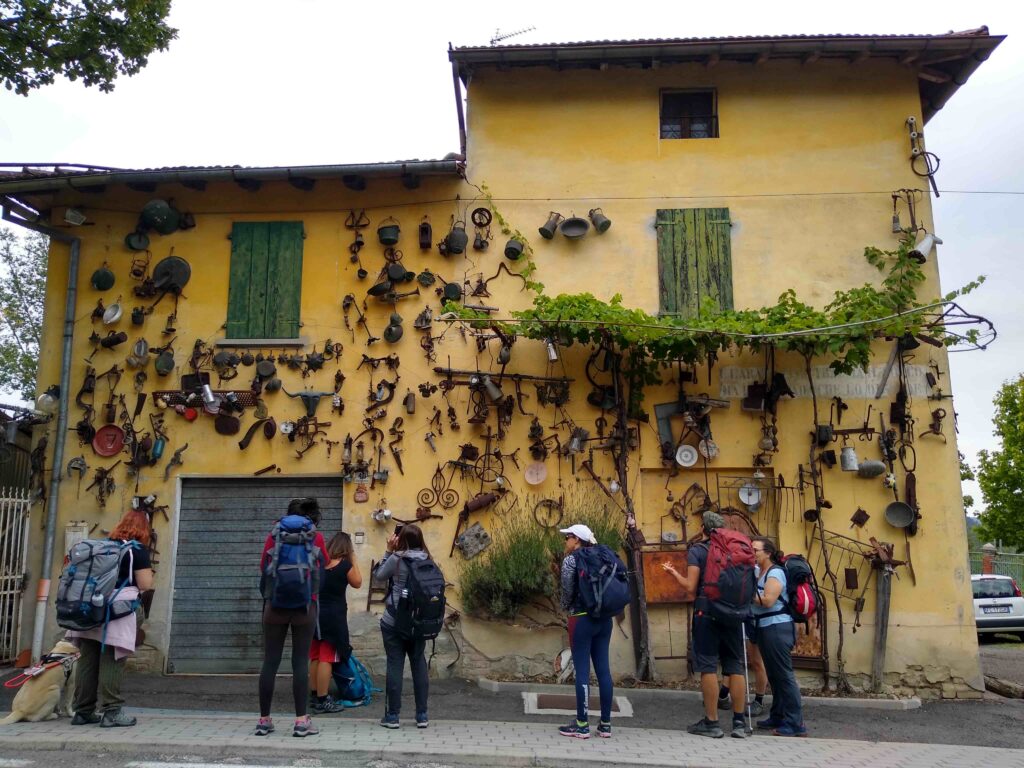 Un gruppo di camminatori di fronte ad una casa di montagna intonacata, la cui facciata è ricoperta da antichi ed arrugginiti oggetti di uso quotidiano, appesi a vari chiodi.