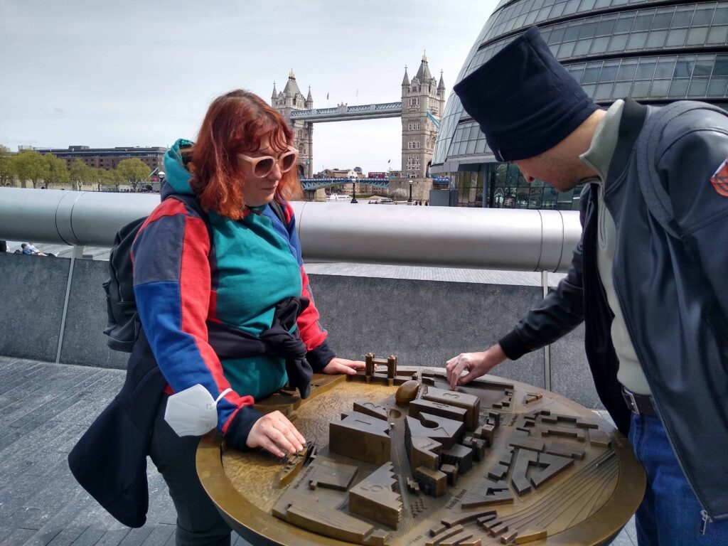 Due viaggiatori toccano un plastico della zona circostante, la piazza del municipio di Londra. Sullo sfondo si vedono le vetrate del futuristico municipio e, in mezzo, le caratteristiche due torri del Tower Bridge.