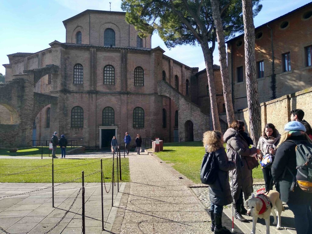 In primo piano, un gruppo di viaggiatori con cane guida. Sullo sfondo, in fondo ad un vialetto, si staglia la basilica di San Vitale a Ravenna, spoglia, angolare e in mattoni rossi.