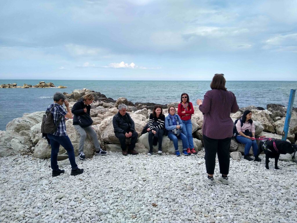 Un'accompagnatrice de La Girobussola, di spalle, parla ad un gruppo di viaggiatori seduti su alcune rocce, su una spiaggia di pietre bianche. Dietro di loro, azzurro e limpido si estende l'Adriatico.