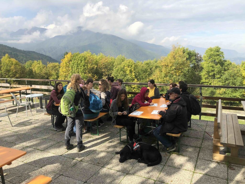 Un gruppo di escursionisti e di mediatori, con e senza disabilità visiva, ascolta una guida seduti intorno a tavoli di legno su un piazzale all'aperto. Davanti a loro hanno delle mappe tattili, mentre oltre la balaustra si vedono le chiome degli alberi e, sul fondo, le cime degli Appennini coperte di nuvole.