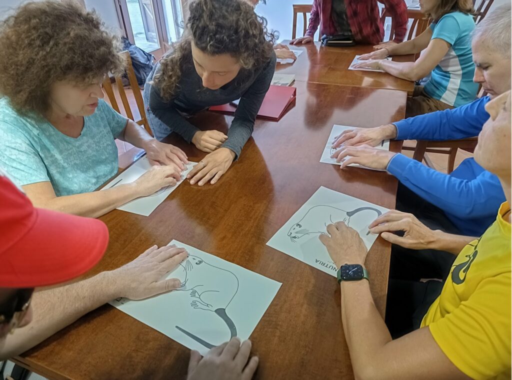 Sedute intorno ad un tavolo, con una guida ad illustrare il disegno, un gruppo di persone esplorano una stampa a rilievo raffigurante una nutria.