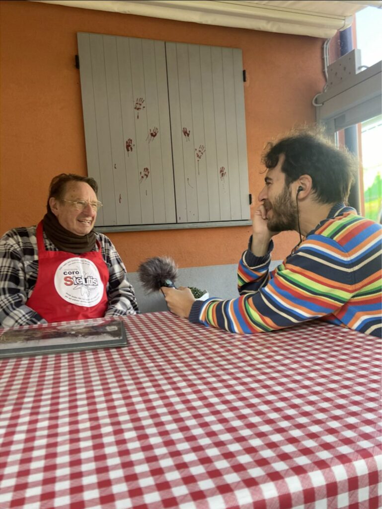 Due persone sedute ad un tavolo, quella più anziana (Fausto Carpani, associazione Ponte della Bionda), sulla sinistra, è intervistata da quella sulla destra con un piccolo registratore portatile a mano.