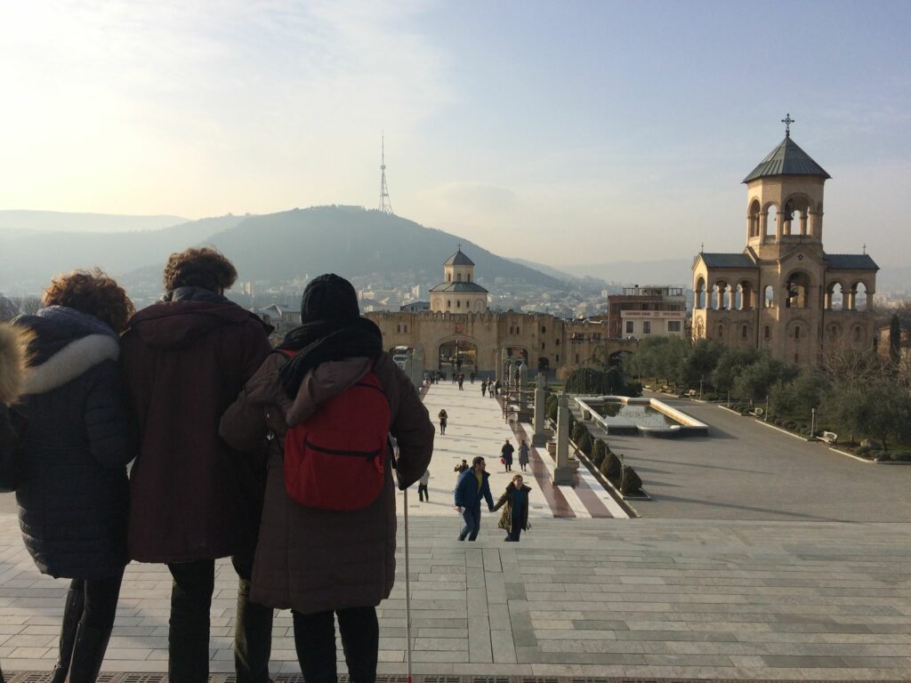 Due viaggiatrici ed un accompagnatore di spalle su una piazza sopraelevata di Tbilisi, sullo sfondo le mura antiche della città, una chiesa paleocristiana e una montagna nella foschia.