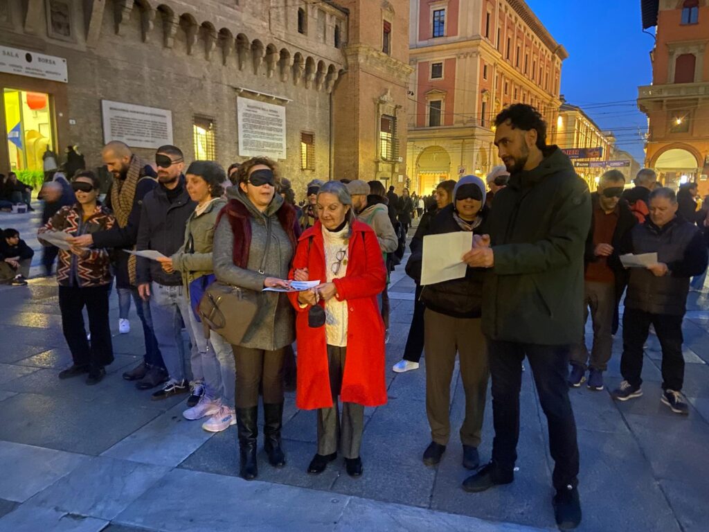 Un gruppo di persone si trovano in Piazza Maggiore a Bologna, davanti all'ingresso della Sala Borsa. Il gruppo è diviso per coppie. Per ogni coppia c'è una persona bendata che esplora la mappa tattile della piazza mentre l'altra persona la guida. 